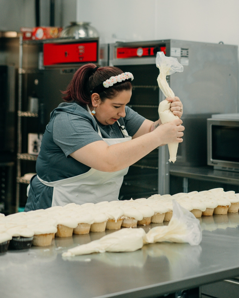 Bridgette Anderson piping frosting onto a long row of cupcakes in a commercial kitchen. She holds a large piping bag and carefully decorates each cupcake on a stainless steel worktable.