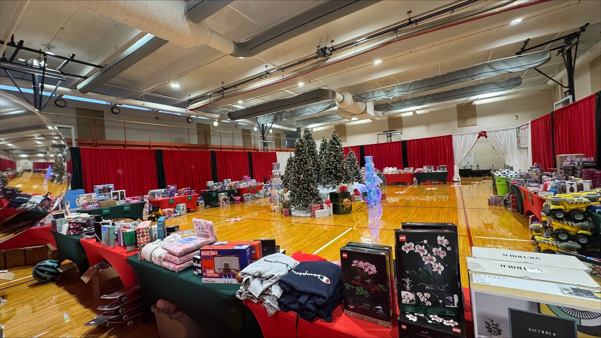 A festive indoor display featuring holiday-themed decorations and gift items arranged on tables in a gymnasium. The setting includes red curtains, Christmas trees, and various boxed products. The polished wooden floor and overhead lighting add to the bright and organized atmosphere.