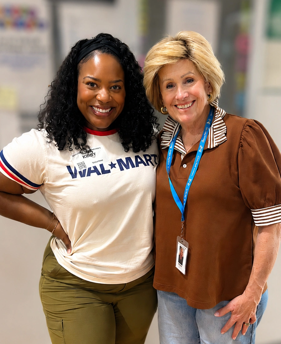 Two women stand side by side, smiling warmly at the camera in an indoor setting. One woman wears a white T-shirt with bold text and casual pants, while the other wears a brown blouse with a striped collar and a blue lanyard with an ID badge. The background is softly blurred, suggesting an office or workplace environment with posters or notices on the walls. The overall mood is friendly, professional, and welcoming.