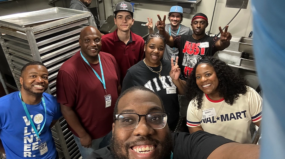 A group of volunteers pose together for a selfie in a commercial kitchen. Metal racks and trays are visible in the background, suggesting a food service or community event setting. The people are smiling, making peace signs, and wearing casual clothing with volunteer name tags. The atmosphere appears friendly, collaborative, and energetic.