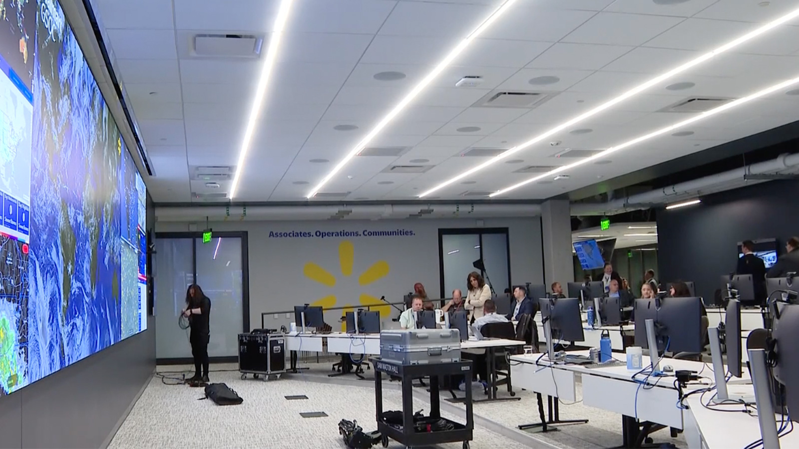 Walmart's Global Security Operations Center, featuring a large digital display wall showing maps and data. The room is equipped with multiple workstations and people collaborating at several workstations. The visible text on the wall reads 'Associates. Operations. Communities.'