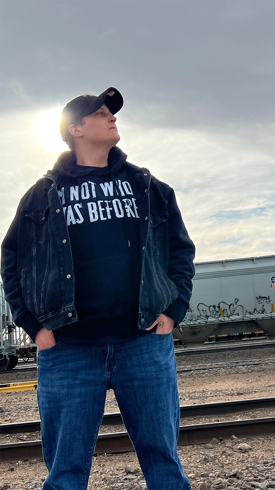 Jayko the ARkiTekk stands by train tracks at sunset, looking to the side.