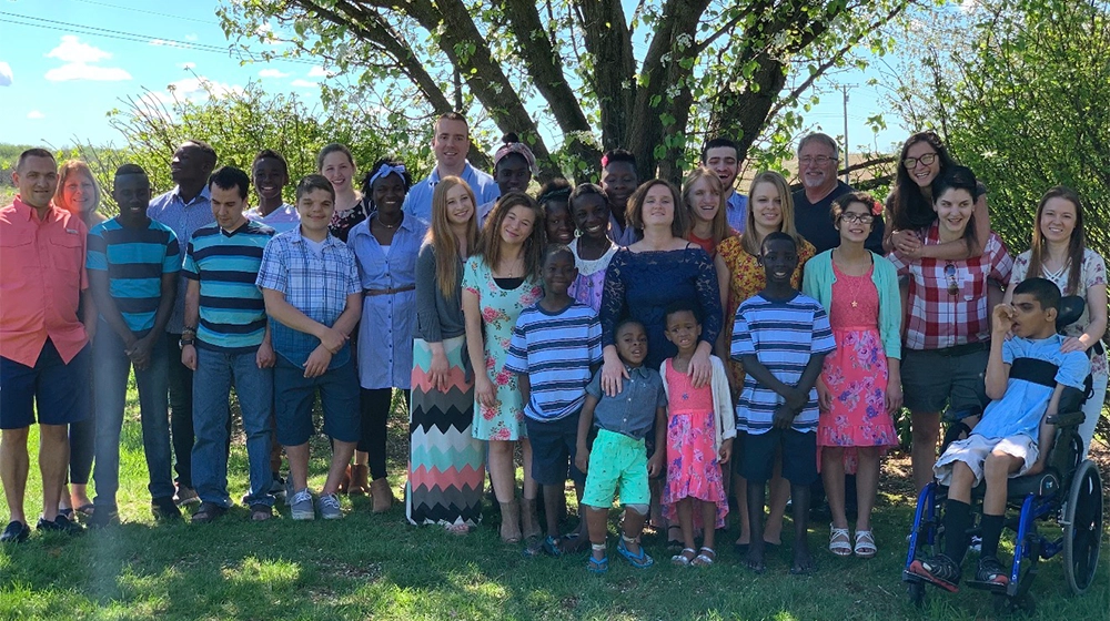 A diverse group of adults and children pose together in front of a leafy tree on a sunny day. The group includes people of various ages, genders, and backgrounds, with some children standing and one child in a wheelchair. The setting is a grassy outdoor area, and the mood is cheerful and relaxed, with bright clothing and natural light. No visible text or numbers are present in the image.