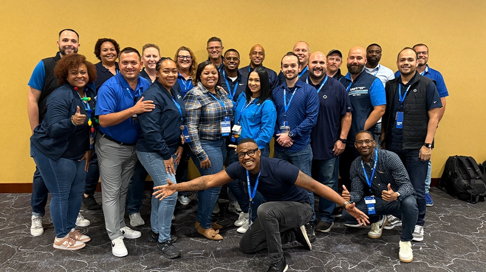 A large group of adults is gathered indoors, posing together in front of a plain yellow wall. Most individuals are wearing blue shirts and conference lanyards, suggesting a professional or team-building event. The mood is upbeat and friendly, with several people smiling and making gestures. The setting appears to be a conference or seminar room with a carpeted floor. No visible numbers or explicit product branding are present in the image.