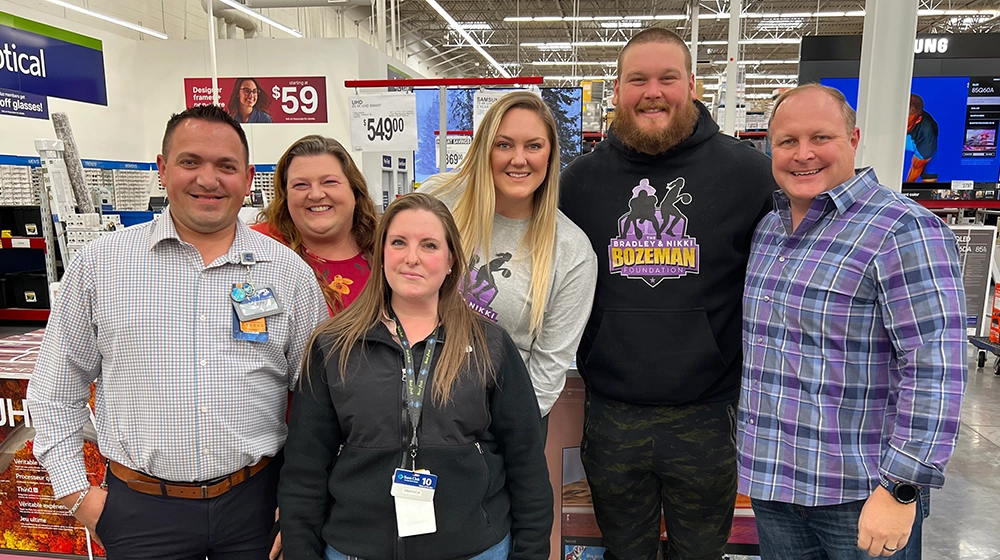 A group of six adults stands together in a brightly lit retail store aisle. The setting features visible price signage, electronics displays, and store uniforms, indicating a workplace or casual shopping environment. One person wears a sweatshirt with the text 'BOZEMAN' and a graphic, while another has a visible employee badge. The mood is casual and friendly, with a mix of plaid and casual attire. The numbers '$59' and '$549.00' are clearly visible on overhead signage.