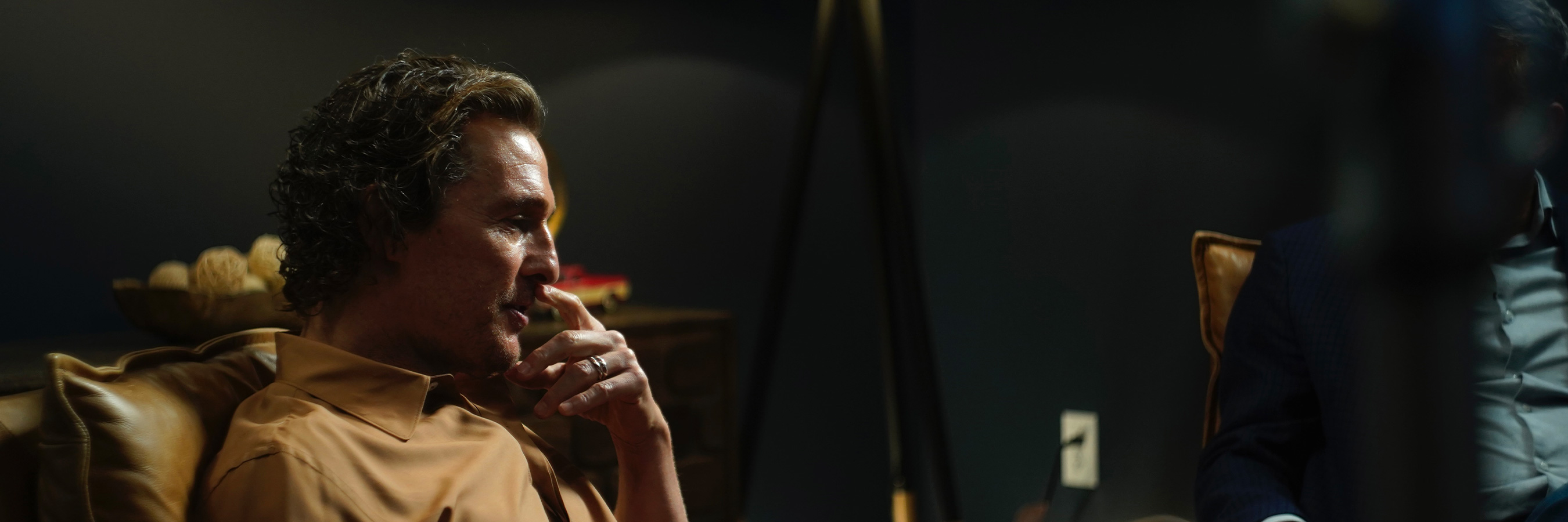 Matthew McConaughey  is seated in a comfortable chair in a dimly lit room, creating a cozy and contemplative atmosphere. A modern floor lamp provides soft lighting, enhancing the warm tones of the setting.