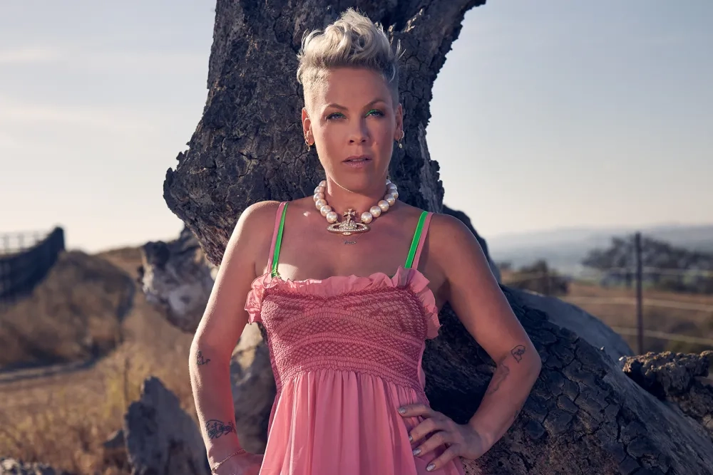 Grammy winner P!NK poses in front of a thick tree trunk, wearing a chunky white necklace and a pink top. She looks directly at the camera.