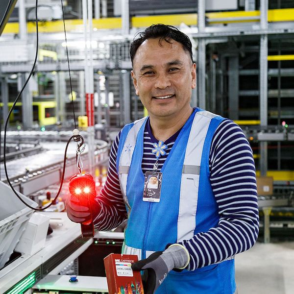 Walmart associate Dal is in a warehouse environment and is holding a red scanner device near a conveyor belt. The setting includes industrial shelving and machinery in the background. The individual is wearing a blue safety vest and gloves. The scanner emits a visible red light, and the worker is handling a rectangular box.