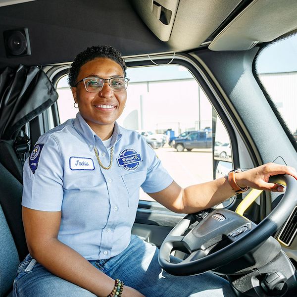 Walmart driver Jackie is seated in the cabin of a truck, wearing a light blue uniform shirt with visible patches and a name tag reading Jackie. The setting appears to be a sunny day. The interior of the truck cabin is well-lit, showcasing the steering wheel.